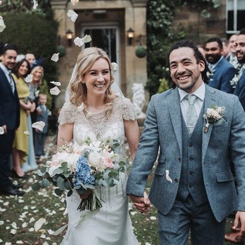 A bride and groom walk hand in hand outdoors in Northumberland, smiling, surrounded by guests throwing beautiful wedding flowers during a joyful celebration.