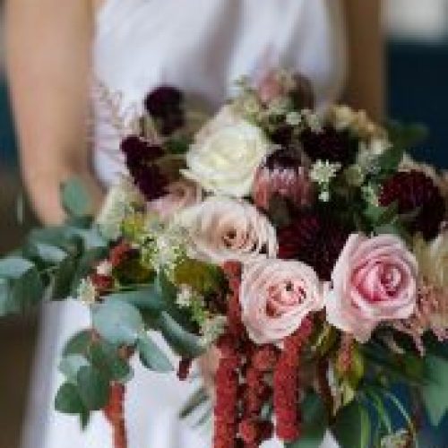 A person in a white dress holds a bouquet of pink, white, and dark red flowers with greenery and trailing burgundy accents—perfect for elegant wedding styling in Newcastle or Northumberland.