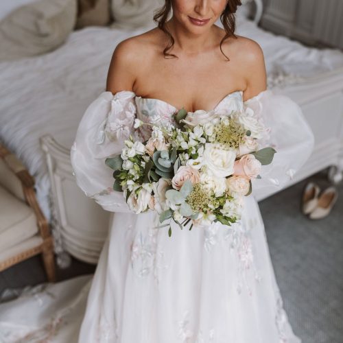 A bride in an off-shoulder white gown holds a stunning Beamish Hall Wedding flowers bridal bouquet of white and pale pink blooms, standing in a bedroom with a bed and a pair of shoes visible in the background.
