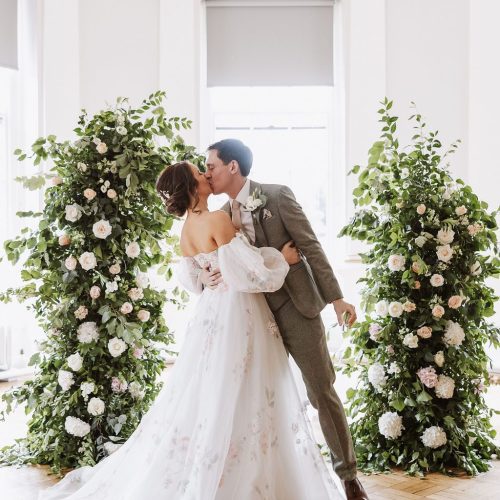 A bride and groom share a kiss indoors during their ceremony, surrounded by tall Beamish Hall Wedding flowers with white and pink blooms and lush greenery.