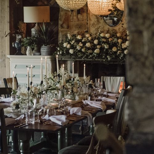 A dining table is set with The Woodmans Arms wedding flowers, candles, glassware, and napkins in an elegant room decorated with greenery and hanging globe lights.
