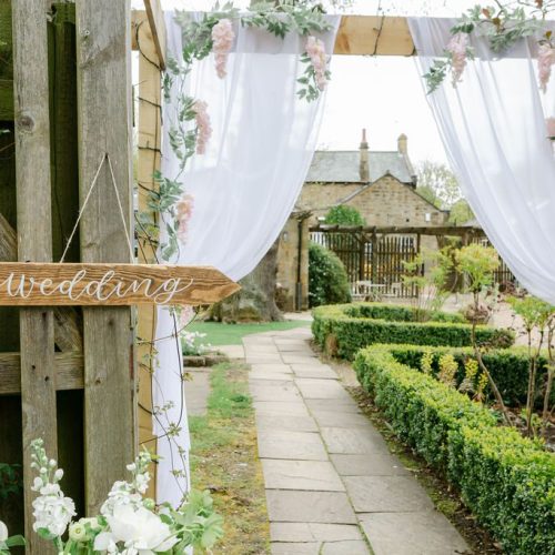 A wooden sign reading "wedding" points toward a garden path lined with greenery and white drapes, leading to an outdoor venue filled with beautiful wedding flowers from a talented Northumberland wedding florist.