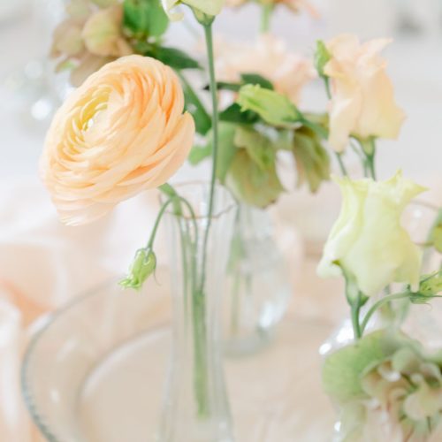 A clear glass vase holds pale peach and light green spring wedding flowers, creating an elegant Northumberland wedding centrepiece on a glass plate with a soft, light background.