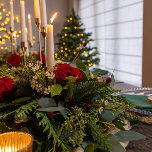 A close-up of a festive dining table styled by n Alexander Weddings features candles, red roses, a greenery centerpiece, and elegant place settings; a decorated Christmas tree and string lights glow in the background.