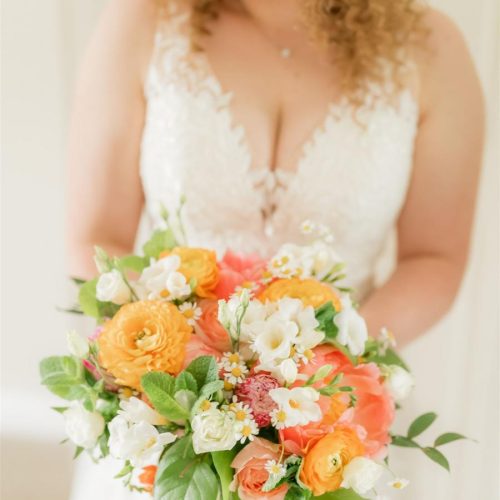 A bride in a white lace dress holds a bouquet of orange, white, and pink wedding flowers with green leaves from a Northumberland florist, smiling and facing slightly to the side.