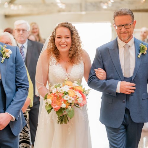 A bride in a white dress holds a bouquet of wedding flowers and walks down the aisle arm-in-arm with an older man, as smiling guests look on at this Northumberland celebration.