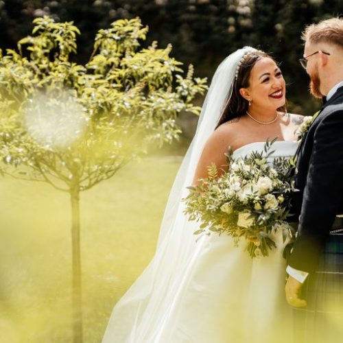 A bride and groom stand outdoors at Shotton Grange in Northumberland, smiling at each other. The bride holds a bouquet of wedding flowers, and both are dressed in wedding attire with lush greenery in the background.