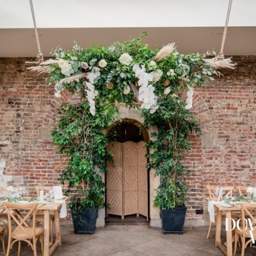 Indoor venue at Middleton Lodge’s Fig House with exposed brick wall, lush wedding flowers, greenery and a floral arch over the doorway, wooden chairs, and tables set with white linen and simple place settings.