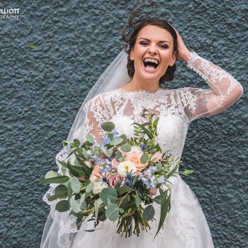 A bride in a lace wedding dress holds a bouquet of flowers and smiles widely while standing against a textured grey wall at Le Petit Chateau in Northumberland.
