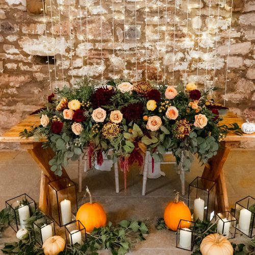A rustic wooden table at Eden Barn, North Yorkshire, is decorated with autumn wedding ceremony flowers, greenery, pumpkins, white candles in glass holders, and a brick wall with string lights in the background.