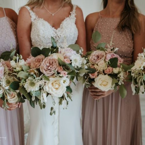 Four women in light dresses, including a bride in a white gown, hold bouquets of white and pink wedding flowers with greenery, crafted by a talented Northumberland florist.