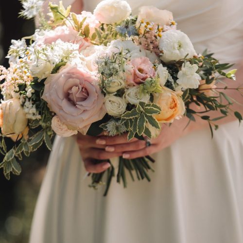 A person in an off-shoulder white dress holds a Spring Bridal Bouquet of pastel roses, peonies, and greenery—perfectly arranged by a wedding florist at Healey Barn.