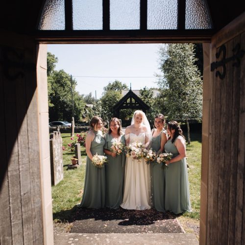 A bride in a white dress stands with four bridesmaids in green dresses outside a chapel, holding bouquets, viewed through an open doorway.