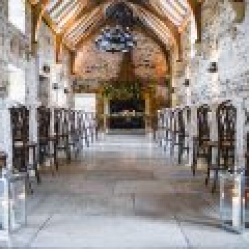 A stone-walled hall at Healey Barn set up for a wedding ceremony, with rows of chairs adorned with white fabric and lanterns lining the aisle.