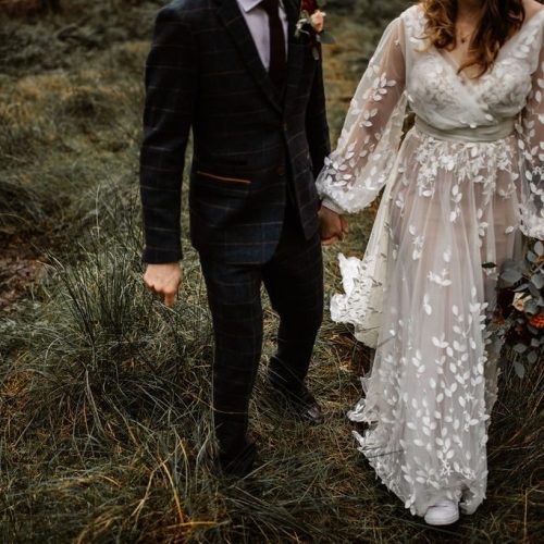 A bride in a white floral dress and a groom in a dark suit walk hand in hand on grass, with the bride holding stunning wedding flowers arranged by a talented Northumberland florist.